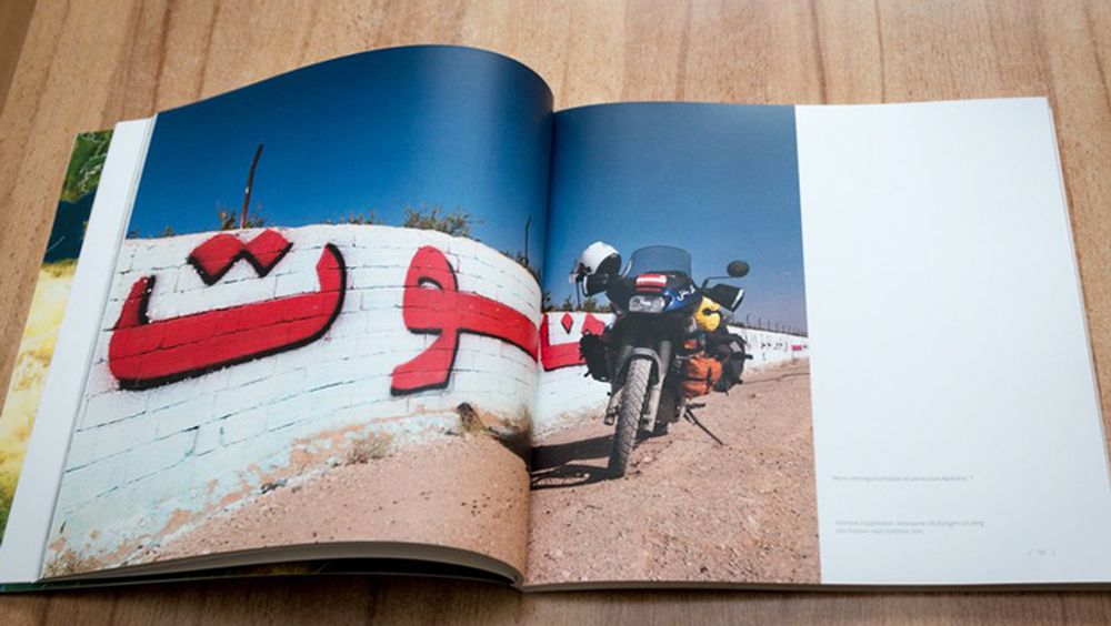 Open book on wooden table. Book shows a large photograph of a loaded motorbike in front of a white wall with persian letters. Deep blue sky.