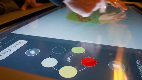 An interactive surface showing a digital color palette and a drawing canvas. Fingers drawing on the canvas.