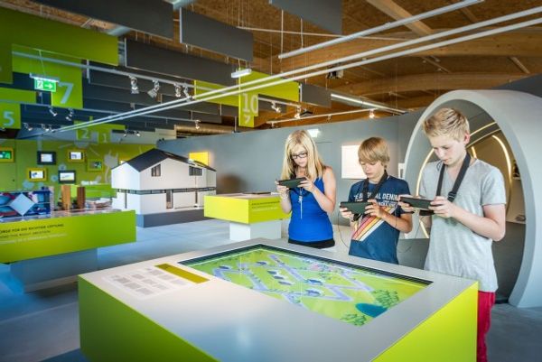 Three children in a museum next to a large display on a table. Each kids holds a device in their hands.